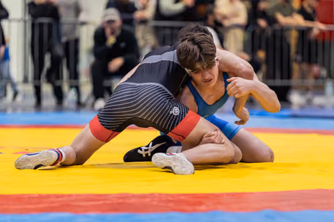 Young wrestler in blue singlet grimaces with focus while controlling opponent in black striped singlet on yellow mat.