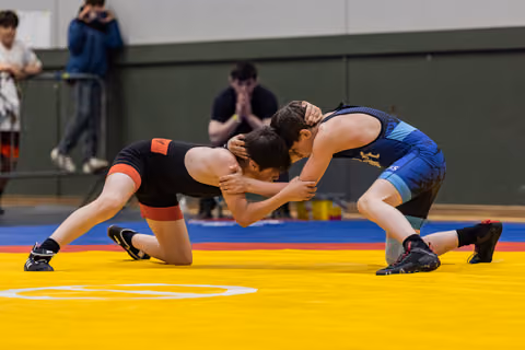 Two young wrestlers in black and blue singlets clinch heads on a yellow mat, spectators watching intently behind.