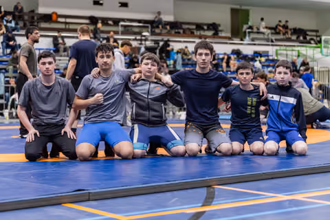 Six young male wrestlers kneel together on a blue mat, arms around each other, posing confidently for a team photo.
