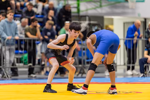 Two young wrestlers in black and blue singlets crouch in ready stances, focused and tense, on a yellow competition mat.