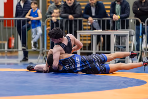 Athlete in black singlet pins opponent in blue 'Mussati' singlet to orange mat, crowd watching behind barriers.