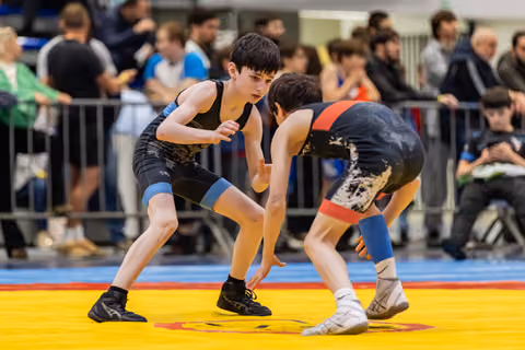 Two young wrestlers in focused stance face each other on a yellow mat, crowd watching in background.