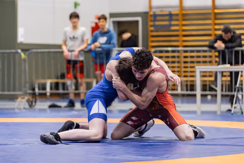 Two young wrestlers locked in an intense clinch on the blue mat, both straining with focused determination during a match.