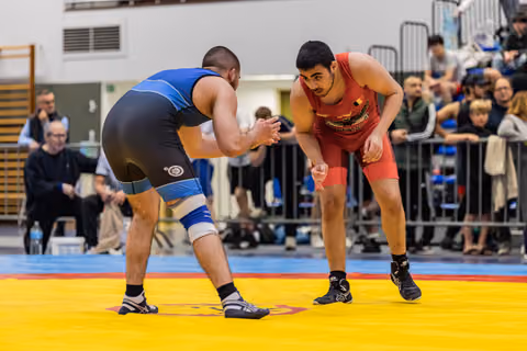 Two wrestlers in blue and red singlets square off on a yellow mat, both crouched and focused, crowd watching behind barriers.