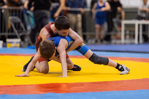 Two young wrestlers grapple on a yellow mat, one in blue controlling the other in red from above.
