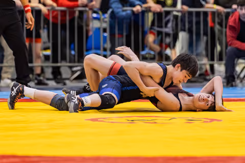 Young wrestler in blue singlet pins opponent to the mat, both showing intense focus during a youth match.