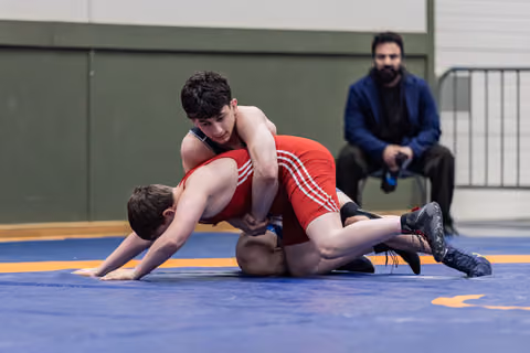 Young wrestler in red dominates opponent on blue mat, controlling from above while coach watches from background.