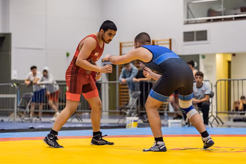 Two wrestlers in red and blue singlets square off on a yellow mat in an indoor sports hall, crowds watching.
