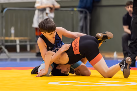 Young wrestler in blue singlet pins opponent to yellow mat, focused expression, indoor sports hall.