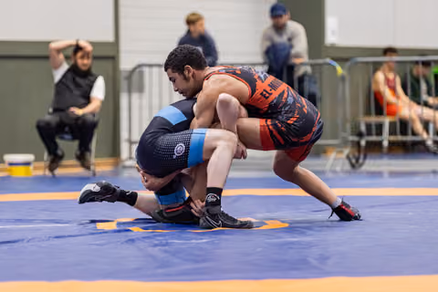 Young wrestler in red singlet drives opponent toward the mat in a powerful double-leg takedown during a match.