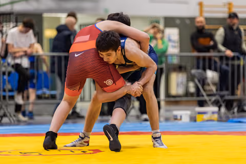 Young wrestler in red singlet locks hands around opponent in blue, both bent low in intense grapple on yellow mat.