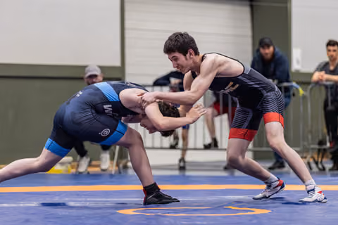 Young wrestler in dark singlet aggressively drives opponent's head down during intense match on blue mat.