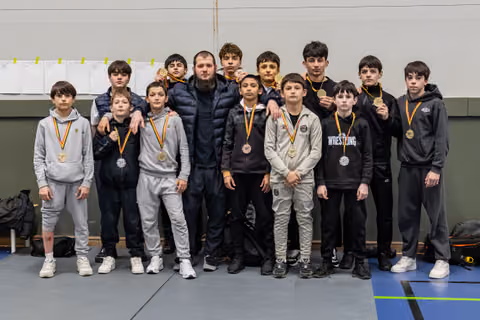 Young wrestlers proudly display gold medals with their coach in a gymnasium after a successful tournament.