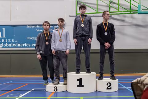 Four young male wrestlers stand on a podium, wearing medals, with the gold medalist standing tallest on position 1.