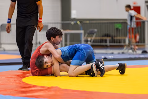 Boy in blue singlet pins opponent in red to the yellow mat while referee watches nearby in gym.