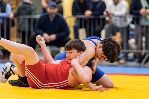 Young wrestler in red grimaces while being pinned by opponent in blue on a yellow competition mat.