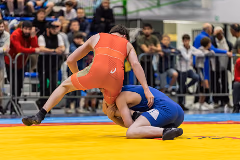 Wrestler in red singlet lifts and drives blue opponent down onto the yellow mat during intense match.