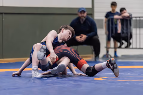 Boy in blue singlet drives opponent to the mat with focused intensity during indoor wrestling match.