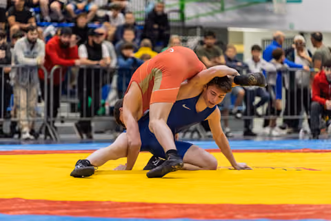 Wrestler in blue singlet braces against opponent in red attempting a takedown on a yellow competition mat.