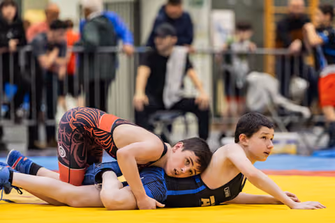 Two young wrestlers compete on a yellow mat, one pinning the other down, both showing intense focus and determination.