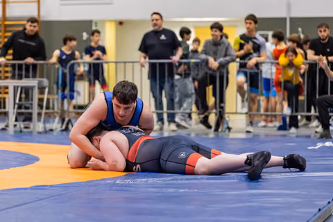 Young wrestler in blue singlet pins opponent face-down on mat as crowd watches intently in background.