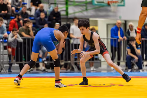 Two young wrestlers in blue and black singlets crouch in an intense standoff on a yellow competition mat, crowd watching.
