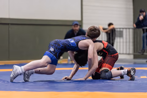 Young wrestler in blue singlet drives forward aggressively while opponent in black and red struggles on the mat.