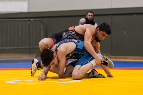 Wrestler in black tiger singlet dominates opponent on yellow mat, both focused and intense during indoor bout.