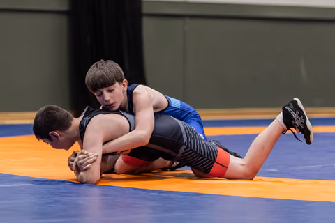 Young wrestler in blue singlet applies a tight body lock on his opponent, pressing him down on the orange mat.