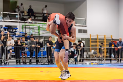 Young wrestler lifts opponent off the mat with intense focus, crowd watching in the background.