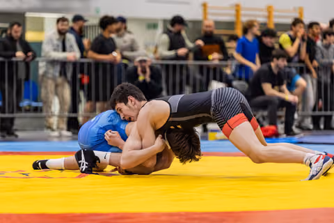 Wrestler in black singlet aggressively drives blue-clad opponent toward the mat, crowd watching intently in background.