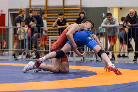Two young wrestlers in red and blue singlets battle on the mat as spectators watch from behind barriers.