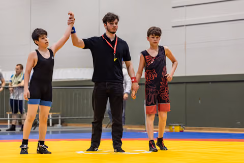 Referee raises young wrestler's hand in victory while defeated opponent stands dejected on yellow mat.