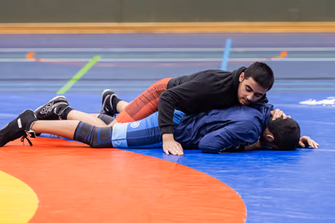 Young wrestler in black top pins opponent in blue singlet to the mat, maintaining dominant top position.