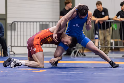 Wrestler in red singlet bent low, grimacing, as blue-suited opponent grips his head with dominant control on blue mat.