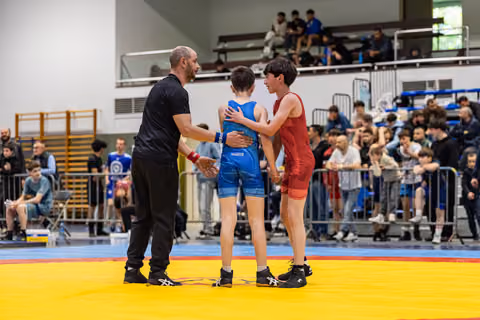 Coach and referee shake hands beside two young wrestlers in blue and red singlets after a bout on the mat.