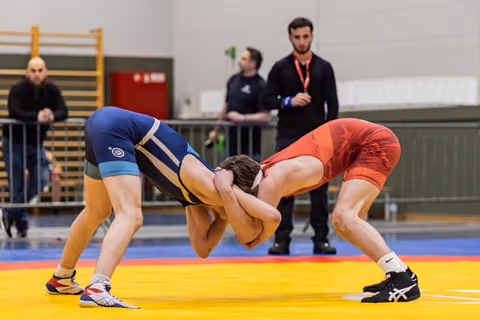 Two wrestlers in blue and red singlets locked in an intense clinch on a yellow mat, referee watching closely.