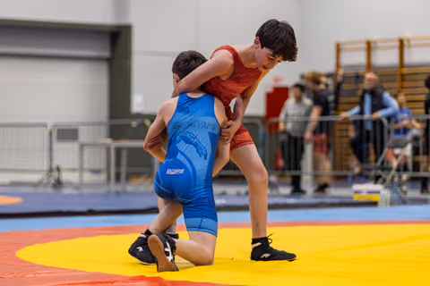 Young wrestler in red singlet dominates opponent in blue, who is down on one knee on a yellow and red mat.