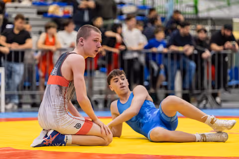 Young wrestler kneels confidently on yellow mat while opponent lies sprawled behind him, crowd watching in background.