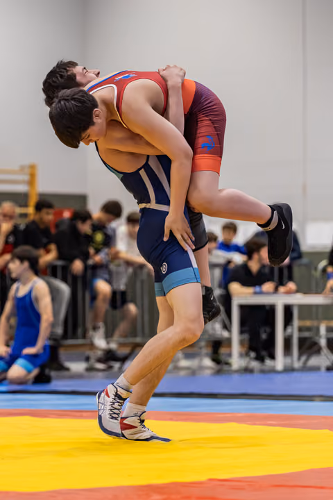 Young wrestler in navy singlet lifts opponent clean off the mat in a powerful body-lock during an indoor tournament.