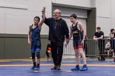 Referee raises the arm of a young wrestler in blue singlet declaring victory, as the opponent stands beside them.