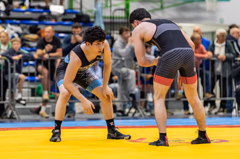 Two young wrestlers in black singlets face off in a crouched, tense stance on a yellow mat before a crowd.