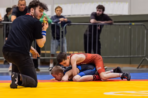 Young wrestler in red singlet pins opponent on mat while referee kneels close, watching intently in indoor sports hall.