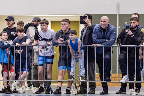 Coaches, athletes, and parents lean on metal barriers, watching intently with phones raised, at an indoor wrestling event.