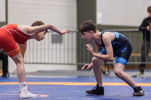 Two young wrestlers in red and blue singlets face off in a crouched stance on the blue mat, focused and ready.