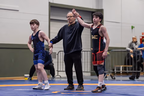 Referee raises the arm of a young wrestler in black-red singlet, declaring victory on a blue mat indoors.
