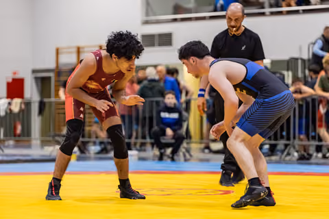 Two young wrestlers in red and blue singlets square off in a tense standoff, referee watching closely behind them.