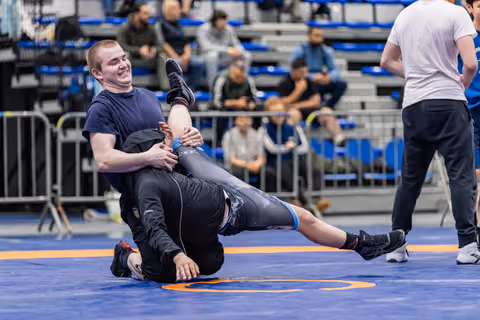 Young wrestler grins while controlling his opponent on the ground during an indoor sports event.