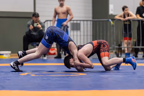 Wrestler in blue singlet drives opponent in red to the mat during an intense youth wrestling bout indoors.
