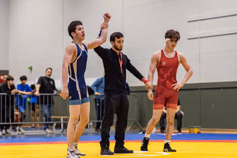 Referee raises the blue singlet wrestler's arm in victory while the red singlet opponent stands deflated nearby.